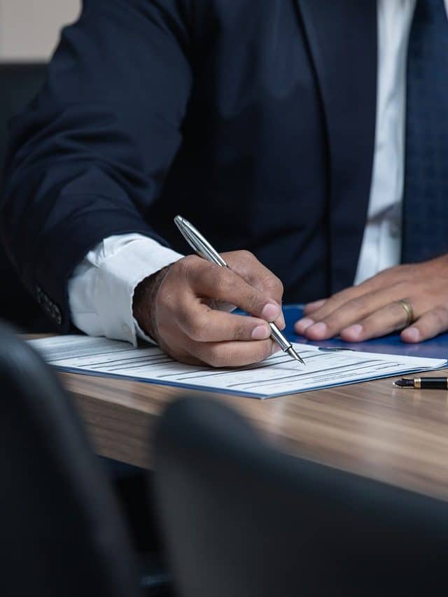 Close-up of a man in a navy suit signing a document at a wooden desk, gold wedding band visible, pen in hand